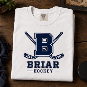 May include: White t-shirt with a navy blue design featuring the letter "B" and crossed hockey sticks. The text "BRIAR HOCKEY" is below the logo. The shirt is folded on a wooden surface.