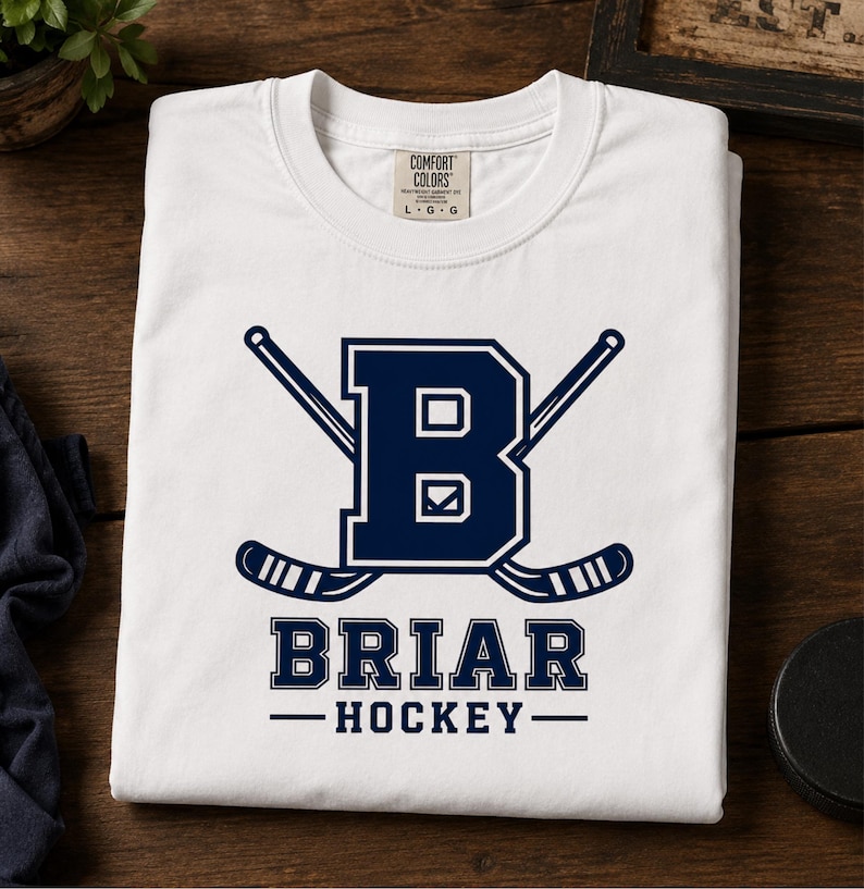 May include: White t-shirt with a navy blue design featuring the letter "B" and crossed hockey sticks. The text "BRIAR HOCKEY" is below the logo. The shirt is folded on a wooden surface.