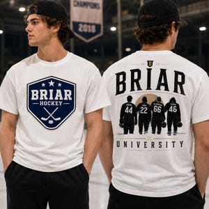 May include: White t-shirt with a navy blue and white hockey-themed graphic on the front. The graphic includes the text "BRIAR HOCKEY" and crossed hockey sticks. The back of the shirt features the word "BRIAR" and silhouettes of hockey players.