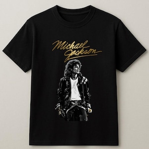 May include: Black t-shirt featuring a gold script of "Michael Jackson" above a black and white image of the singer. The image shows Michael Jackson wearing a black jacket and white shirt.