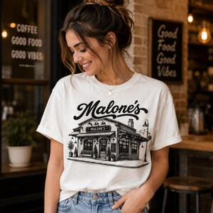 May include: Cream-colored t-shirt featuring a black and white illustration of a building with the text "Malone's" above it. The shirt is being worn by a person in blue jeans.