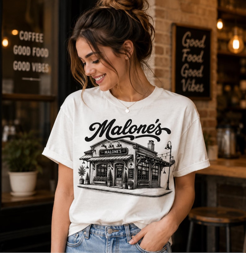 May include: Cream-colored t-shirt featuring a black and white illustration of a building with the text "Malone's" above it. The shirt is being worn by a person in blue jeans.