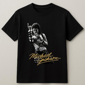 May include: Black t-shirt featuring a black and white image of Michael Jackson performing on stage. The image includes the text "Michael Jackson" in gold script. The shirt is a classic crew neck style.