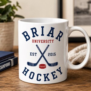 May include: White ceramic mug featuring the text "Briar University Hockey" in navy blue and red. The design includes crossed hockey sticks, a red puck, and the year "2015". The mug has a curved handle and is set on a wooden surface.