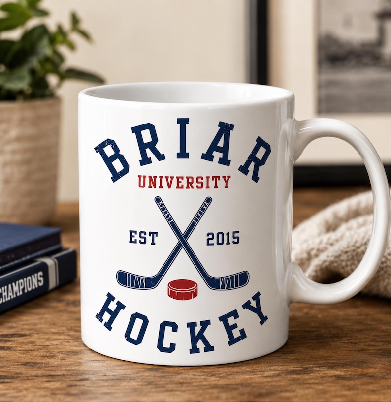 May include: White ceramic mug featuring the text "Briar University Hockey" in navy blue and red. The design includes crossed hockey sticks, a red puck, and the year "2015". The mug has a curved handle and is set on a wooden surface.