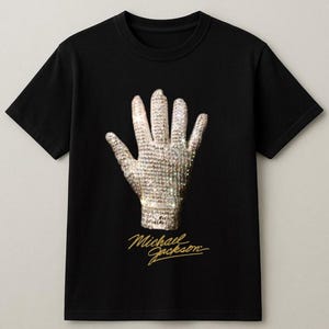 May include: Black t-shirt featuring a sparkling, jeweled glove design. The glove is a prominent image on the shirt, with the name "Michael Jackson" in gold script below. The shirt is a classic crew neck style.