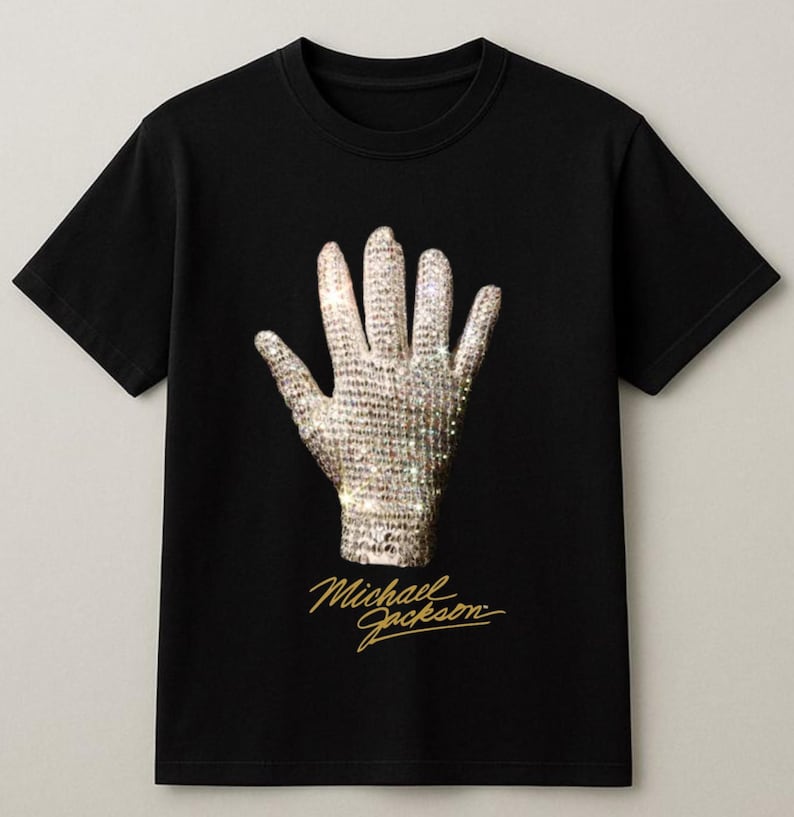 May include: Black t-shirt featuring a sparkling, jeweled glove design. The glove is a prominent image on the shirt, with the name "Michael Jackson" in gold script below. The shirt is a classic crew neck style.