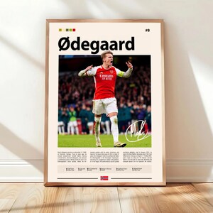 May include: Framed poster featuring a soccer player in a red and white uniform, with the name "Ødegaard" at the top. The poster includes a photo of the player, his signature, and biographical text. The frame is a light brown color.