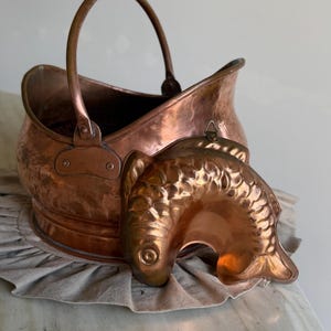 Puede incluir: Un cubo para carbón de cobre con un asa curva y una base remachada. En la parte frontal tiene un molde decorativo de cobre con forma de pez, con escamas y aletas detalladas, que incluye una pequeña anilla metálica para colgarlo.