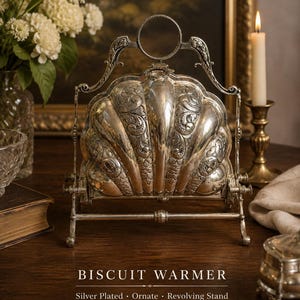 Puede incluir: Un calentador de galletas plateado con un diseño ornamentado en forma de concha y grabados intrincados. El objeto está montado sobre un soporte giratorio con un asa circular en la parte superior. El texto en la imagen dice: BISCUIT WARMER, Silver Plated, Ornate, Revolving Stand, Vintage Elegance for Your Table.