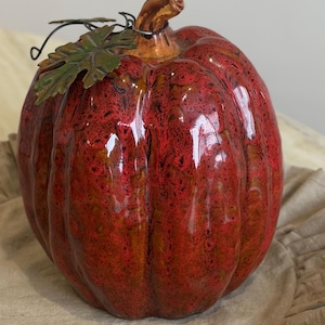 May include: A glossy ceramic pumpkin figurine featuring a deep red and orange mottled glaze. The pumpkin has a prominent brown stem and a decorative dark green metal leaf accent attached with a thin black wire near the top of the gourd.