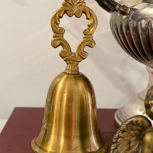 May include: A polished brass hand bell featuring a smooth, flared body and an ornate, openwork handle with symmetrical scrollwork detailing. The bell has a bright, reflective metallic finish and a distinct, flared rim at the base of the structure.