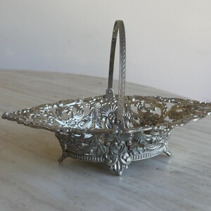 May include: A silver-coloured metal basket featuring an intricate openwork filigree design with ornate scrollwork patterns. The basket has a rectangular base with four small feet and a single upright arched handle attached to the centre sides.
