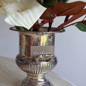 May include: A silver-toned metal urn-shaped vase with a flared rim and ornate handles, containing white calla lilies and dark brown magnolia leaves. A small rectangular tag with the word Archive in white script is attached to the front.