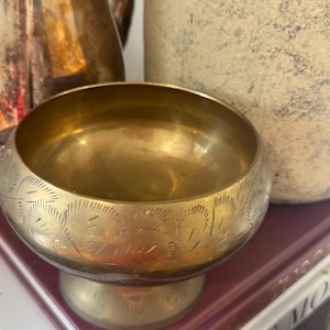 May include: A small, round, gold-coloured brass bowl with a pedestal base. The exterior surface of the bowl features a repeating etched pattern of fan-shaped motifs and linear strokes. The metal has a polished finish with visible metallic sheen.
