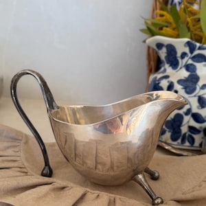 May include: A polished silver gravy boat featuring a curved handle, a pointed pouring spout, and three small ball-tipped feet. The metallic surface reflects the surrounding environment, showing a smooth, rounded body and a wide opening.