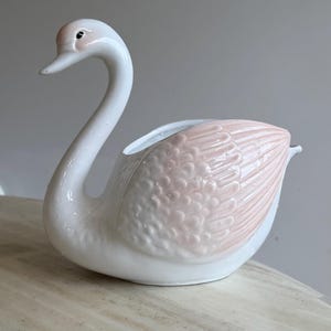 May include: A ceramic swan-shaped planter or dish featuring a glossy white finish on the neck and base, with soft pastel pink detailing on the textured wing feathers. The swan has a curved neck, a small black eye, and an open top cavity.