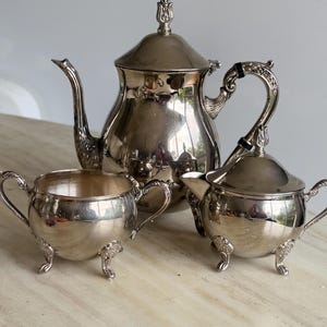 May include: A silver-plated tea set consisting of a teapot with a lid, a sugar bowl, and a creamer. Each piece features a polished metallic finish, curved handles, and decorative feet. The teapot has a tall spout and a finial on the lid.