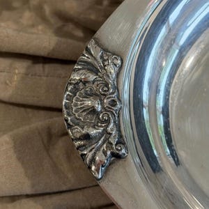 May include: A polished silver-toned metal serving tray featuring a prominent decorative handle. The handle is cast with a detailed relief pattern that includes a central shell motif surrounded by scrolling acanthus leaf designs.