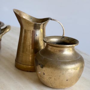 May include: A brass pitcher with a curved handle and a flared spout positioned next to a small, round brass pot with a wide, open rim. Both vessels feature a metallic gold-toned finish with visible signs of natural patina and surface wear.