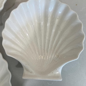 Puede incluir: Un plato de cerámica blanca con forma de concha de vieira, acabado brillante y crestas radiantes. El cuenco presenta una abertura ancha en forma de abanico y una base pequeña y plana en la parte inferior, con un diseño limpio y neutro.