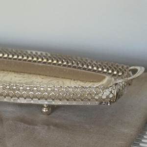 May include: Silver-coloured rectangular tray with a decorative, openwork border and small ball feet. The tray has a patterned interior and small handles on each end. The tray is displayed on a neutral-coloured surface.