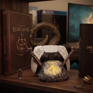 May include: A white and black game controller rests on a textured rock-shaped stand featuring a glowing yellow symbol. The stand is flanked by two sword-like ornaments and a curved, weathered sculpture, positioned between two large books titled Elden Ring.