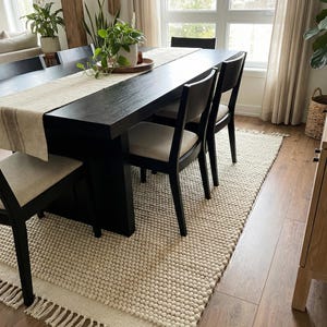 Pode incluir: Uma sala de jantar com uma grande mesa preta e seis cadeiras pretas. Um corredor de mesa bege e plantas em vasos adornam a mesa. Um tapete texturizado creme com franjas fica sob a mesa, em um piso de madeira.
