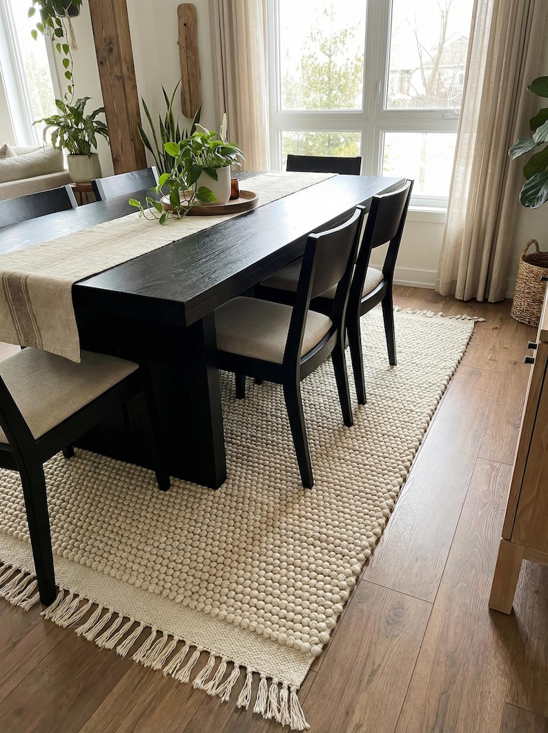 Pode incluir: Uma sala de jantar com uma grande mesa preta e seis cadeiras pretas. Um corredor de mesa bege e plantas em vasos adornam a mesa. Um tapete texturizado creme com franjas fica sob a mesa, em um piso de madeira.