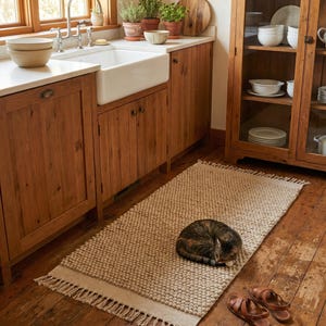 Pode incluir: Um tapete bege texturizado com borda de franjas est&aacute; em um ch&atilde;o de madeira. Um gato enrolado descansa no tapete. Sand&aacute;lias de couro marrom colocadas perto. A cozinha tem arm&aacute;rios de madeira e uma pia branca. O tapete tem cerca de 150 cm de comprimento.