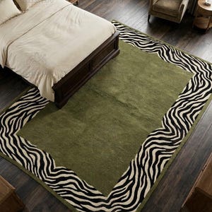 May include: A rectangular area rug featuring a solid olive green centre panel surrounded by a wide border with a black and white zebra stripe pattern. The rug is positioned on a dark wood floor, partially underneath a bed with cream bedding.