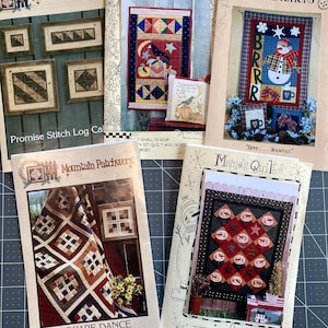 May include: Four quilting pattern books with different designs. The first book is titled "Mountain Patchwork" and features a log cabin design. The second book is titled "Meme's Quilts" and features a quilt with a crow and moon design. The third book is titled "Kalico Kreations" and features a snowman design. The fourth book is titled "Meme's Quilts" and features a snowman design.