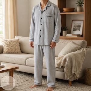 May include: Light grey pyjama set with black trim. The long-sleeved button-down shirt has a chest pocket and black buttons. The matching trousers have a relaxed fit. The pyjamas are made of a soft, textured fabric.