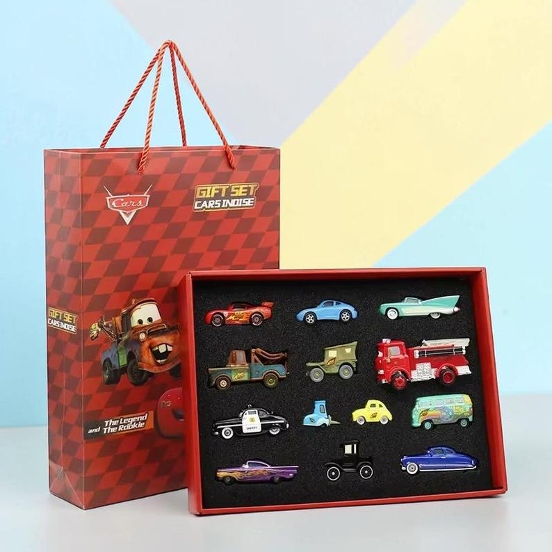 May include: A red gift bag and a red box set featuring miniature car figurines. The gift bag has a Cars theme with a diamond pattern and the text "GIFT SET CARS INOISE". The box contains 12 colourful car models.
