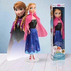 May include: A doll of a character from the movie Frozen, wearing a black top, blue skirt, and pink cape. The doll has blonde hair in braids and is standing on a white stand. The box reads "Fashion".