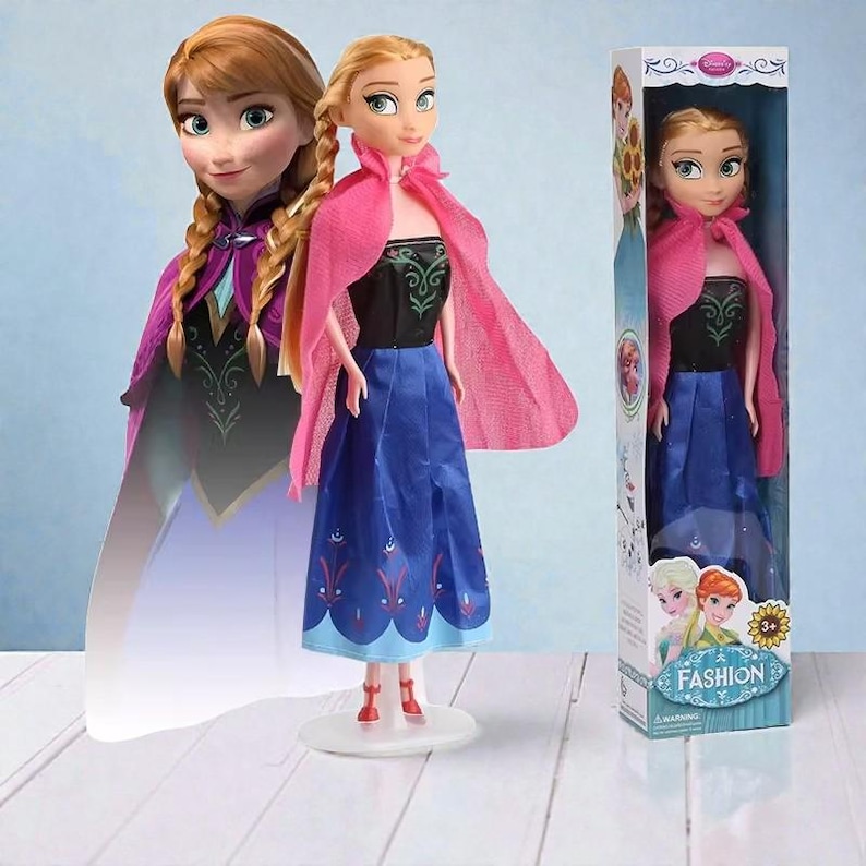 May include: A doll of a character from the movie Frozen, wearing a black top, blue skirt, and pink cape. The doll has blonde hair in braids and is standing on a white stand. The box reads "Fashion".