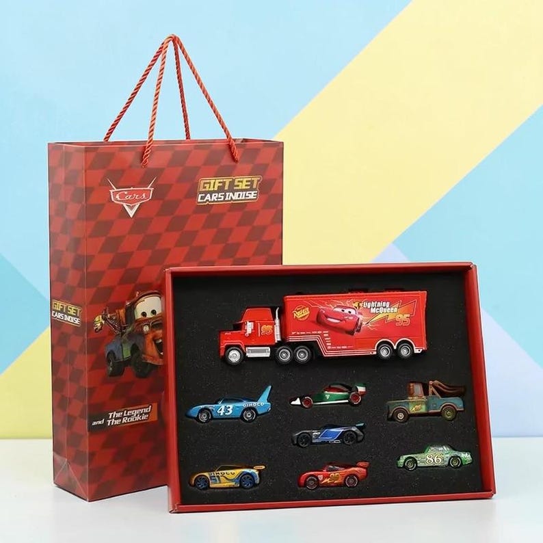 May include: A red gift set featuring toy cars from the film Cars. The set includes a red articulated lorry with a trailer, and several miniature race cars. The gift bag and box have the text "Cars Indise".