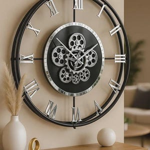 Op de afbeelding: Grote, ronde wandklok met een zwart metalen frame en zilveren Romeinse cijfers. De wijzerplaat is zwart met zichtbare tandwielen. De klok is gemonteerd op een beige muur boven een houten console.