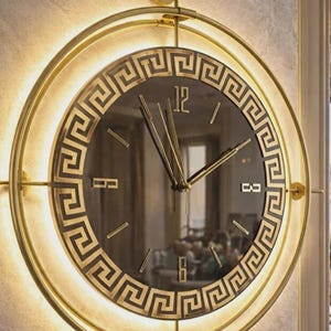 Puede incluir: Un reloj de pared redondo, dorado, con esfera negra y diseño de greca. El reloj tiene manecillas doradas y un marco dorado con un borde iluminado. El reloj está montado en una pared clara y texturizada.