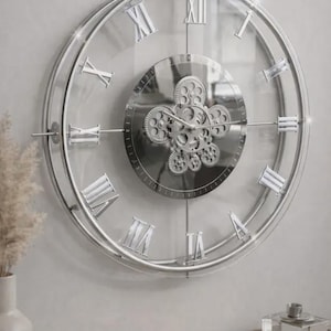May include: A large, round wall clock with a silver metal frame and Roman numeral hour markers. The clock face is clear, revealing the wall behind it. The center features a decorative gear design. The clock is mounted on a light gray wall.