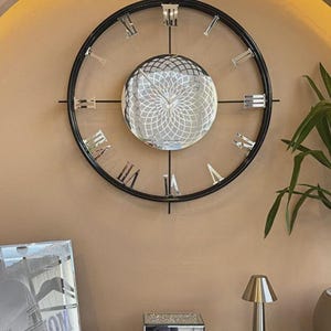 Peut inclure: Une grande horloge murale ronde avec un cadre en métal noir et des chiffres romains. Le cadran est une sphère miroir avec un motif géométrique. L'horloge est fixée sur un mur beige, avec des objets décoratifs en dessous.