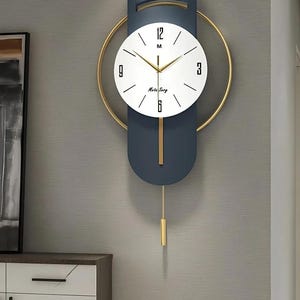 Puede incluir: Reloj de pared moderno con diseño azul marino y dorado. El reloj tiene una esfera blanca con números negros y manecillas doradas. Un péndulo dorado cuelga debajo. El reloj está montado en una pared gris.