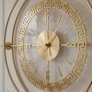 Pode incluir: Um relógio de parede circular com uma moldura de metal dourado e um centro transparente. O mostrador apresenta uma borda com padrão de grega dourada, marcadores de hora dourados e os números 12 e 6 em acabamento dourado com ponteiros.