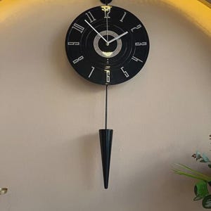 May include: Black wall clock with a pendulum. The clock face is black with white numerals and hands. The pendulum is a black, cone-shaped design. The clock is mounted on a light beige wall.
