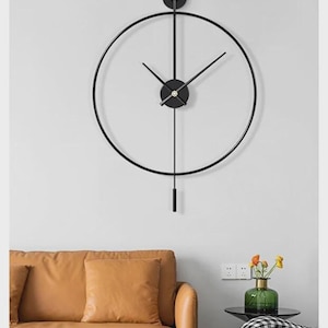Puede incluir: Un reloj de pared moderno y minimalista con marco y manecillas de metal negro. El reloj tiene un diseño circular con un péndulo. El fondo incluye un sofá de cuero marrón, una pequeña mesa auxiliar y un sillón a cuadros.