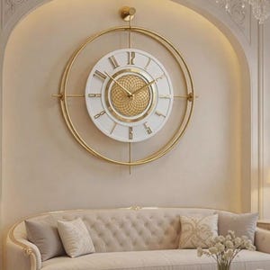Può includere: Un grande orologio da parete con cornice dorata, quadrante bianco e dettagli dorati. L'orologio presenta numeri romani e un centro decorativo. L'orologio è montato su una parete beige sopra un divano beige con cuscini decorativi.