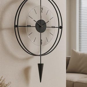 Puede incluir: Un reloj de pared moderno de metal negro con un diseño minimalista. El reloj presenta una esfera de cristal transparente con manecillas e indicadores de hora negros. Un péndulo decorativo cuelga debajo. El reloj está montado en una pared clara.