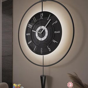 Pode incluir: Um relógio de parede preto moderno com um design circular e um fundo branco iluminado. O relógio apresenta algarismos romanos e ponteiros brancos. Um pêndulo preto pende por baixo. O texto "TASARIM TESCİL BELGESİ" é visível.