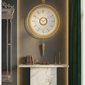 Op de afbeelding: Een goudomrande wandklok met een witte wijzerplaat en gouden cijfers hangt boven een console met marmeren blad. De klok heeft een slinger en is gemonteerd op een donkergrijze muur met een gouden rand. Boeken en decoratieve items staan op de tafel.