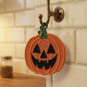 Könnte beinhalten: Ein gestickter Kürbis-Aufnäher in leuchtendem Orange mit einem dunkelgrünen Stiel und einem schwarzen Jack-o'-Lantern-Gesicht mit dreieckigen Augen und gezacktem Mund, der an einem Metallhaken an einer kleinen Schlaufe am Stiel hängt.